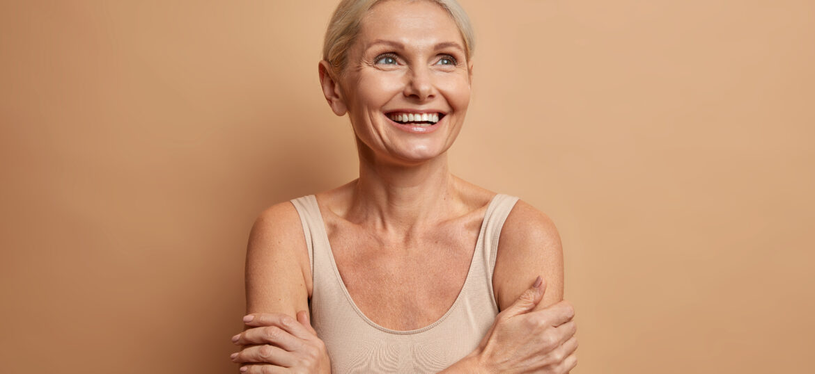 Attractive positive wrinkled fifty years old woman looks gladfully above keeps arms foded has well cared complexion healthy skin white teeth isolated over brown background. Beauty and age concept