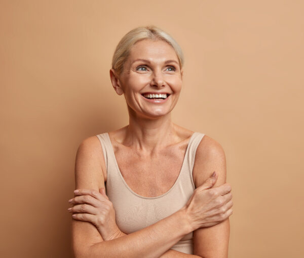 Attractive positive wrinkled fifty years old woman looks gladfully above keeps arms foded has well cared complexion healthy skin white teeth isolated over brown background. Beauty and age concept